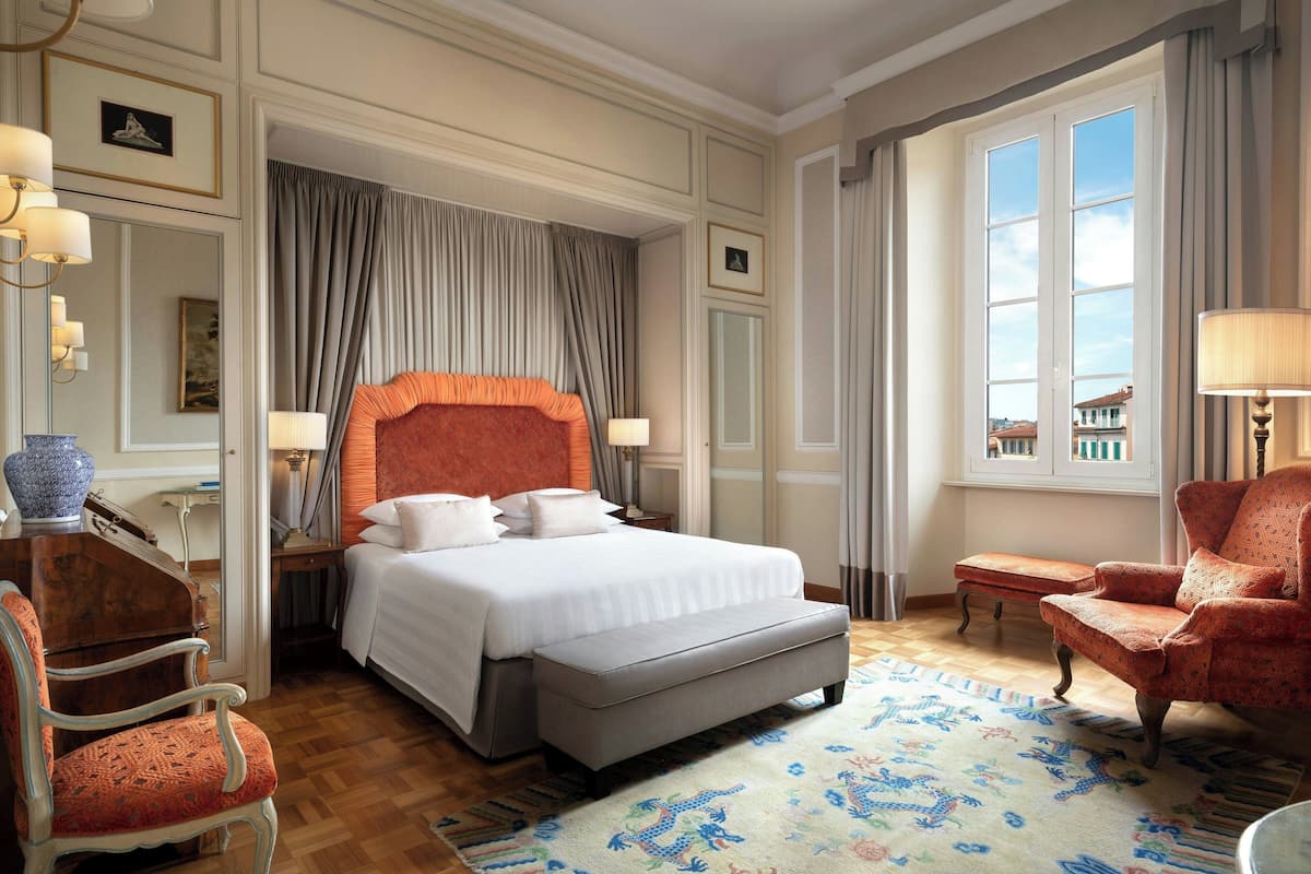 A hotel room with a large bed, a sitting area with a chair, a piano, and a view of buildings outside the window.