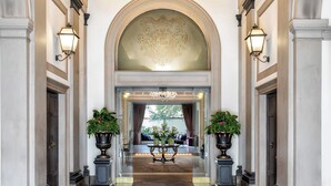 A grand hallway with a high arched ceiling, ornate wall sconces, and a chandelier. There are potted plants on either side of the hallway leading to a seating area with a table and chairs.