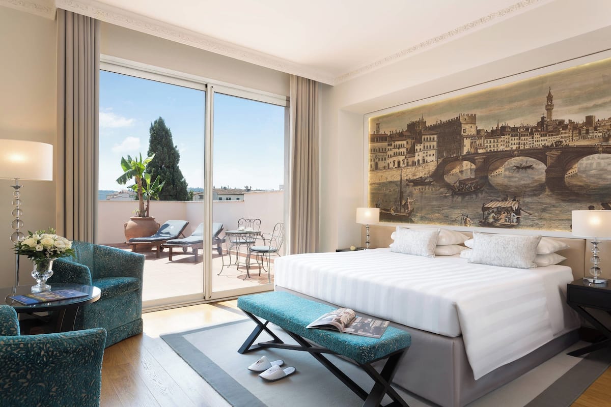 A hotel room with a large bed, a bench, a chair, a table with a vase of flowers, and a painting of a bridge.