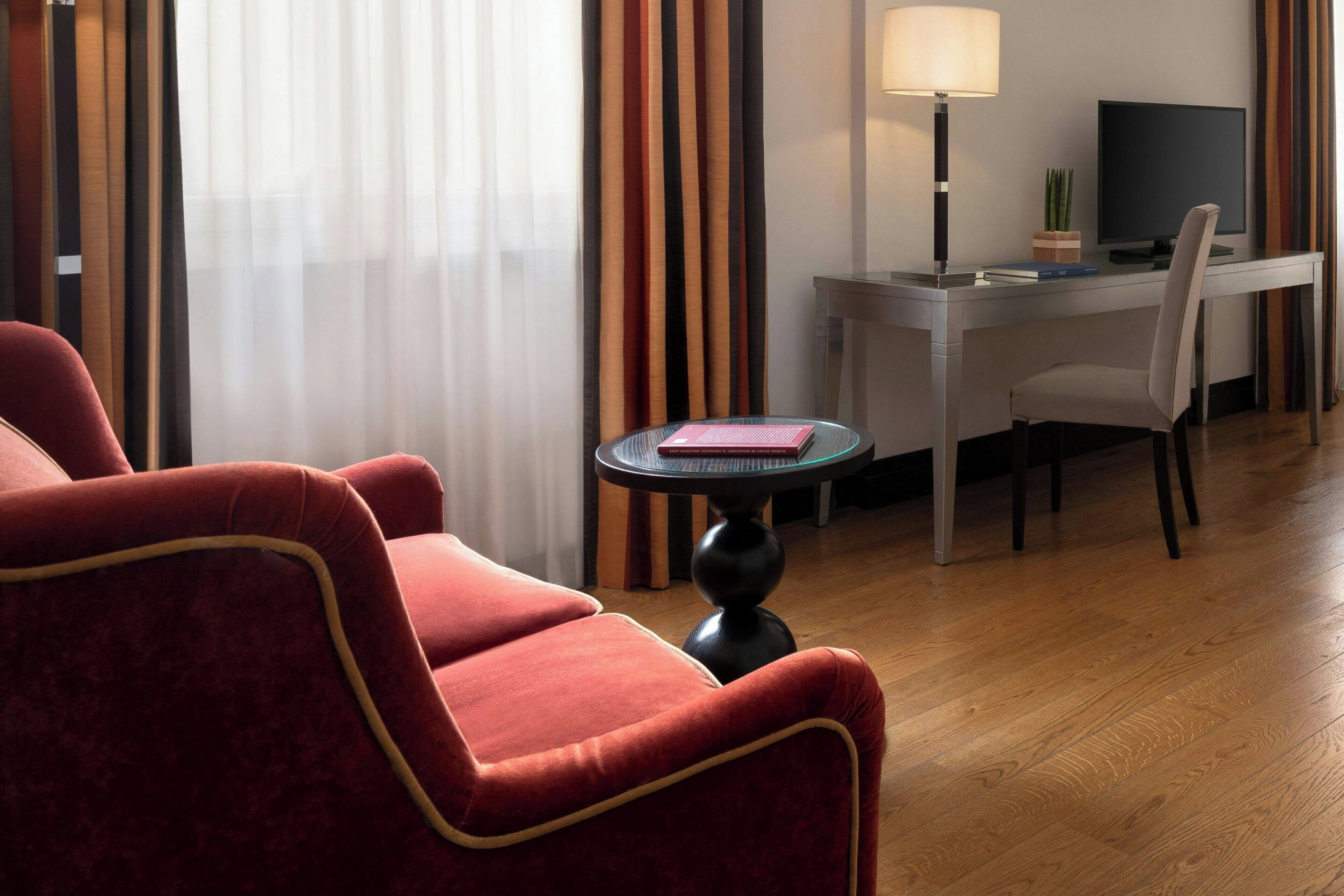 Una camera d'albergo con una poltrona rossa, un tavolino rotondo con un libro, una scrivania con un computer e una lampada.