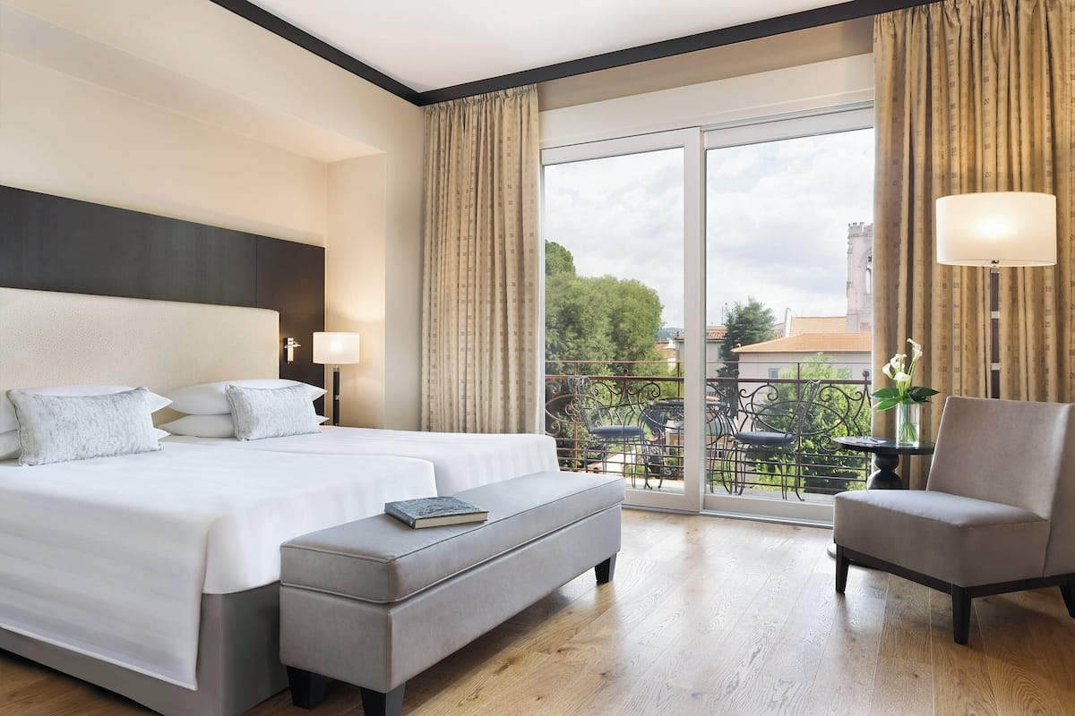 A hotel room with a large bed, a sitting area with a chair, and a balcony with a view of the city.