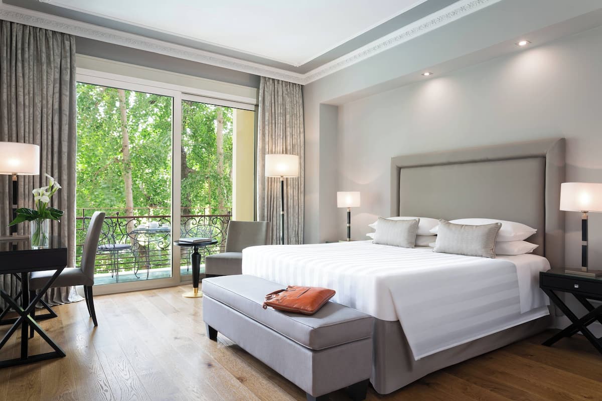 A modern hotel room with a large bed, a desk, and a balcony with a view of trees.