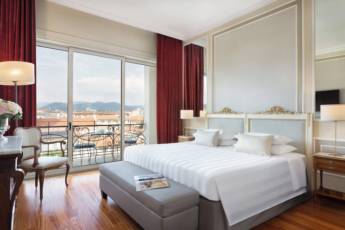 A hotel room with a large bed, a desk, a chair, and a balcony with a view of the city.