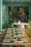 Un restaurant avec une longue table en bois dressée pour le repas, un ordinateur portable posé sur la table et un mur vert orné de plantes en arrière-plan.