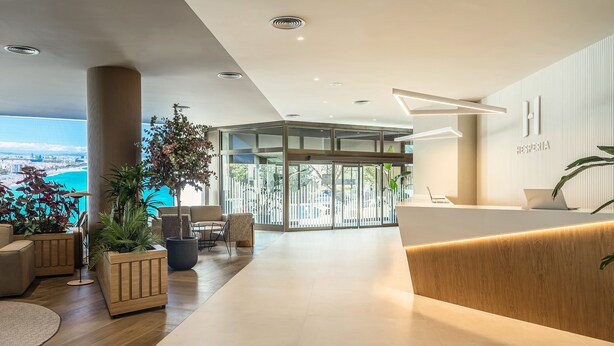 A modern hotel reception area with a wooden reception desk, seating, and large windows offering a city view. - Hesperia Sant Just (Sant Just Desvern)