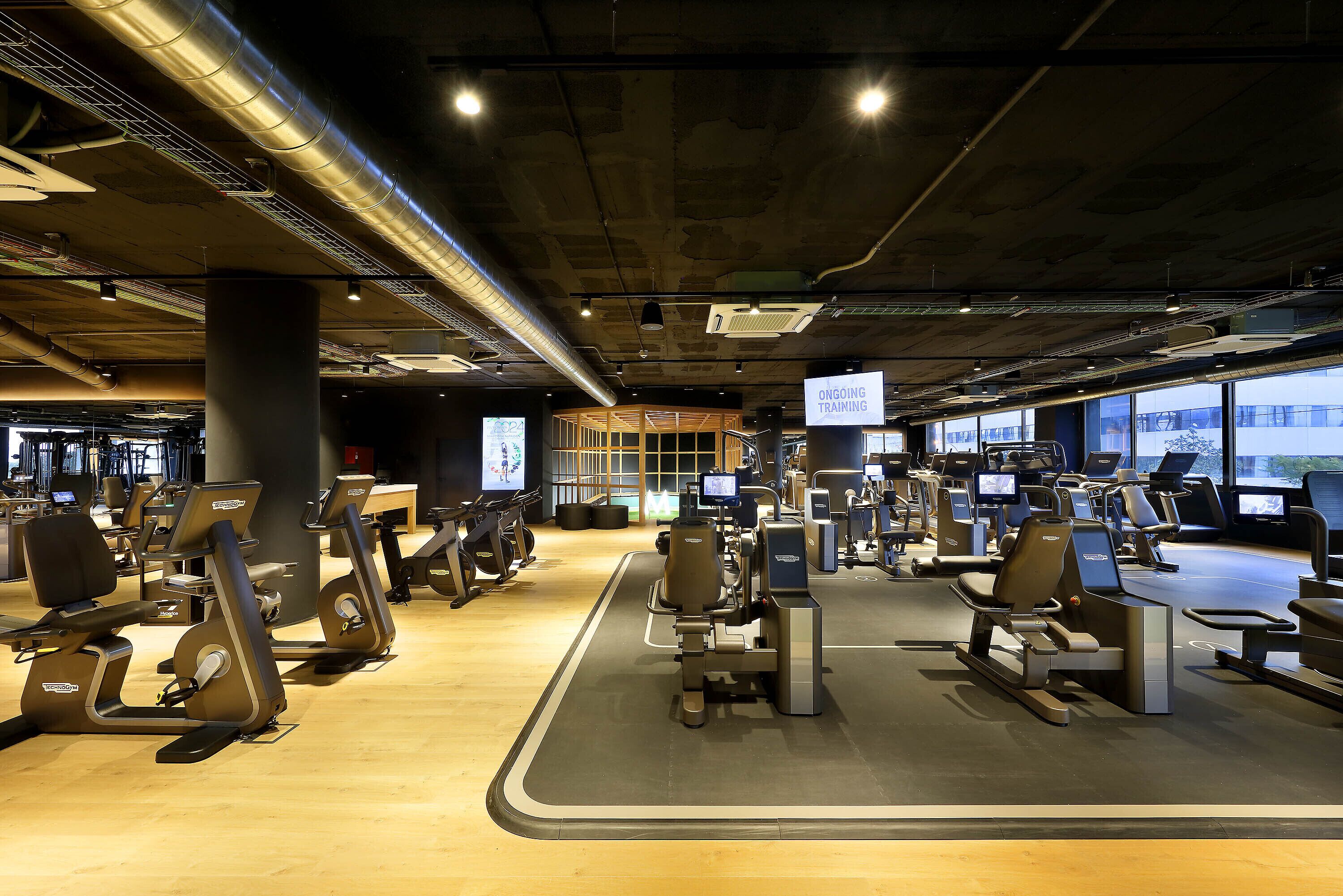 Un gimnasio con diversas máquinas de ejercicio, amplias ventanas y un cartel que indica entrenamiento en curso.