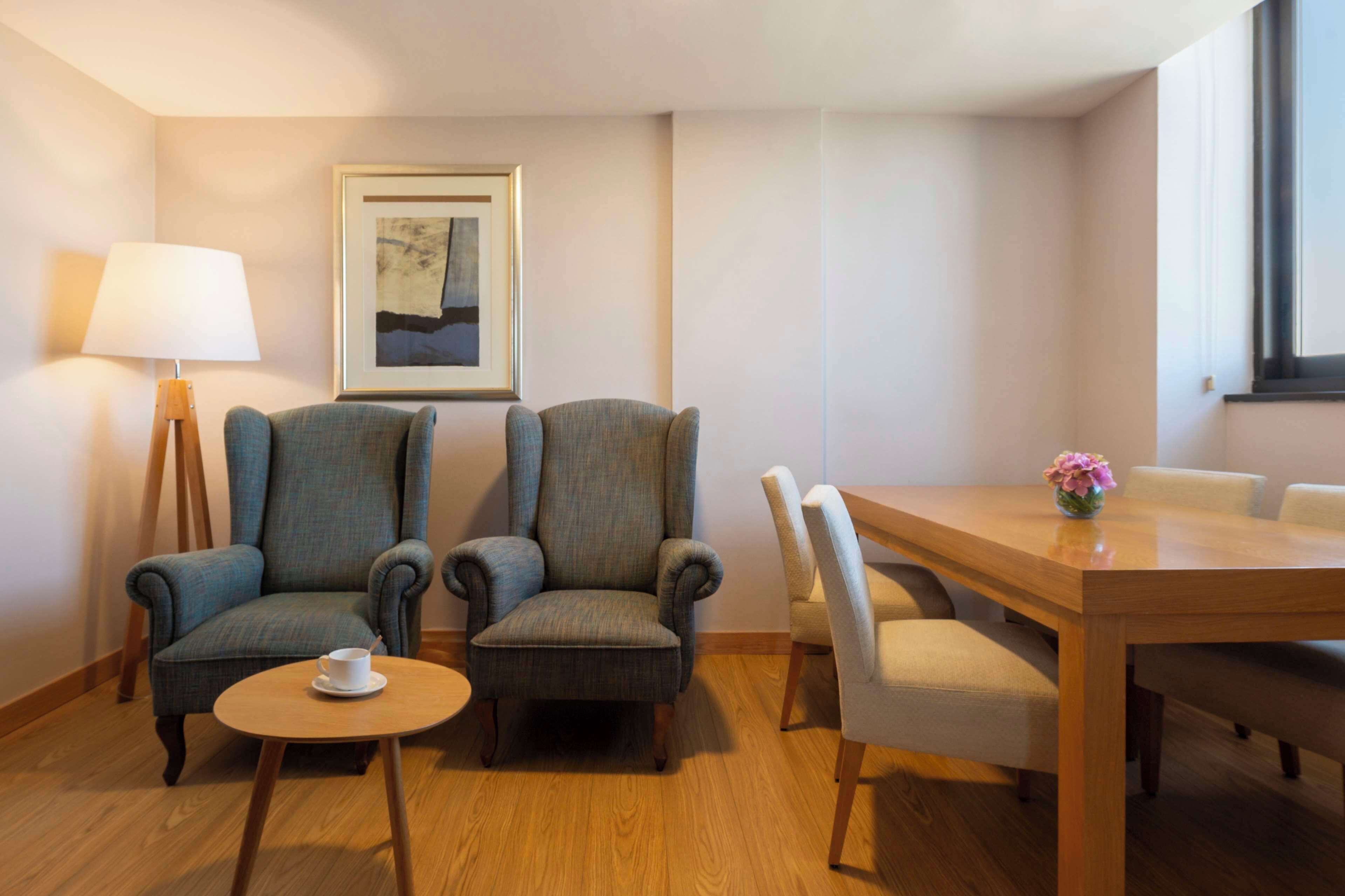 Een hotelkamer met twee fauteuils, een kleine ronde tafel met een kopje, een eettafel met stoelen, een lamp en een lijst met een afbeelding aan de muur.