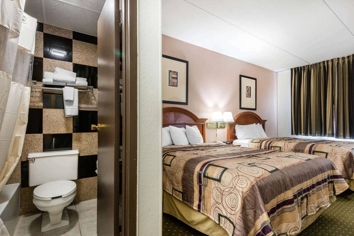A hotel room with a checkered bathroom wall, a toilet, two beds with patterned bedding, a wooden headboard, a bedside table with a lamp, and a framed picture on the wall.