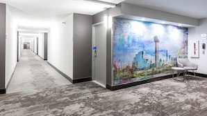 Un couloir d’hôtel avec une fresque représentant un paysage urbain sur le mur, une chaise et une porte avec un panneau indiquant la sortie.