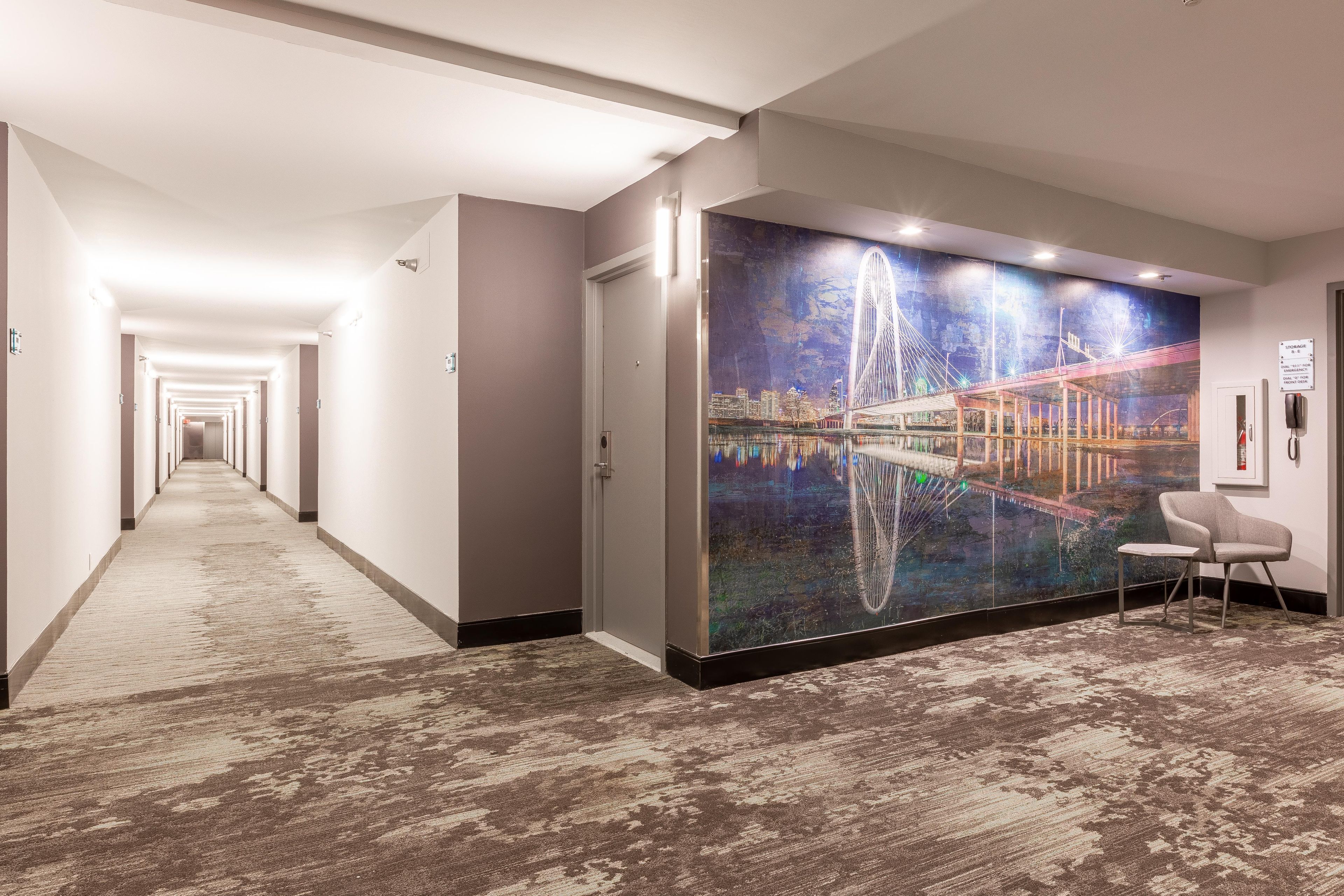 A hotel hallway with a carpeted floor, a large wall mural of a bridge, and a chair.