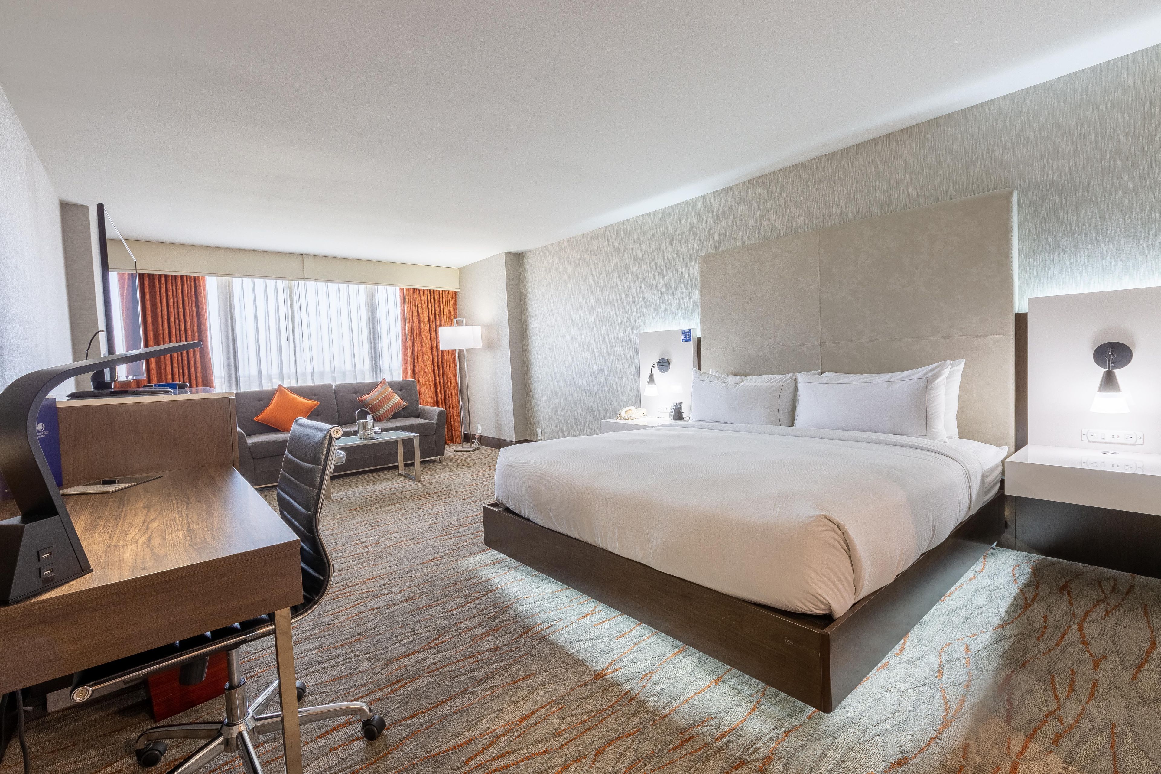 A hotel room with a large bed, a desk with a computer, a sofa, and a window with curtains.