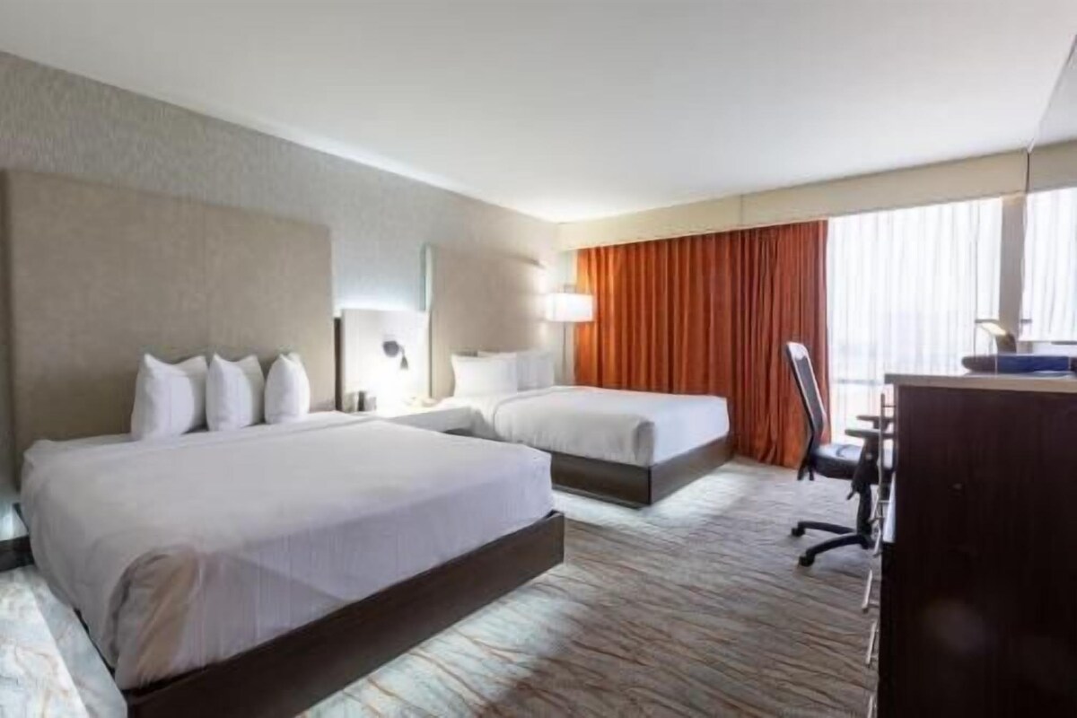 A hotel room with two beds, a desk, a chair, and a large window with curtains.