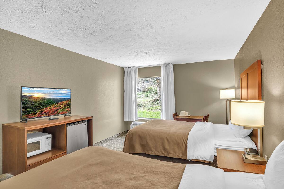 A hotel room with two beds, a TV, a desk with a microwave, and a window with curtains.