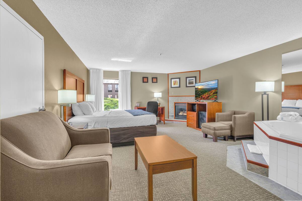 A hotel room with a bed, sofa, armchair, table, and a bathroom with a bathtub.