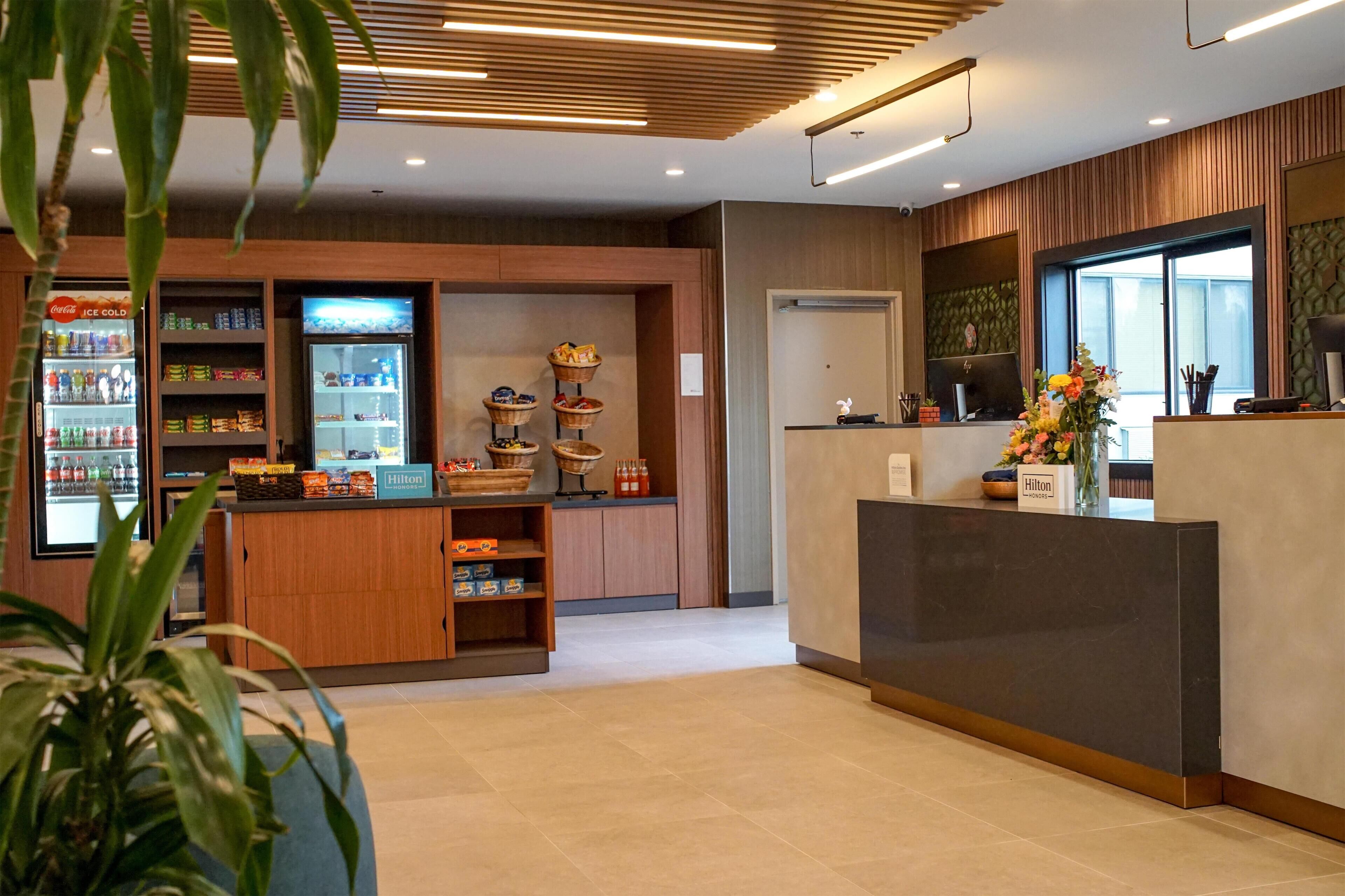 A hotel reception area with a counter, a Coca-Cola refrigerator, snack shelves, and a floral arrangement.