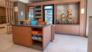 Un espace d’accueil d’hôtel comprenant un comptoir, un réfrigérateur rempli de boissons, des étagères avec des en-cas et un présentoir avec des paniers contenant des collations.