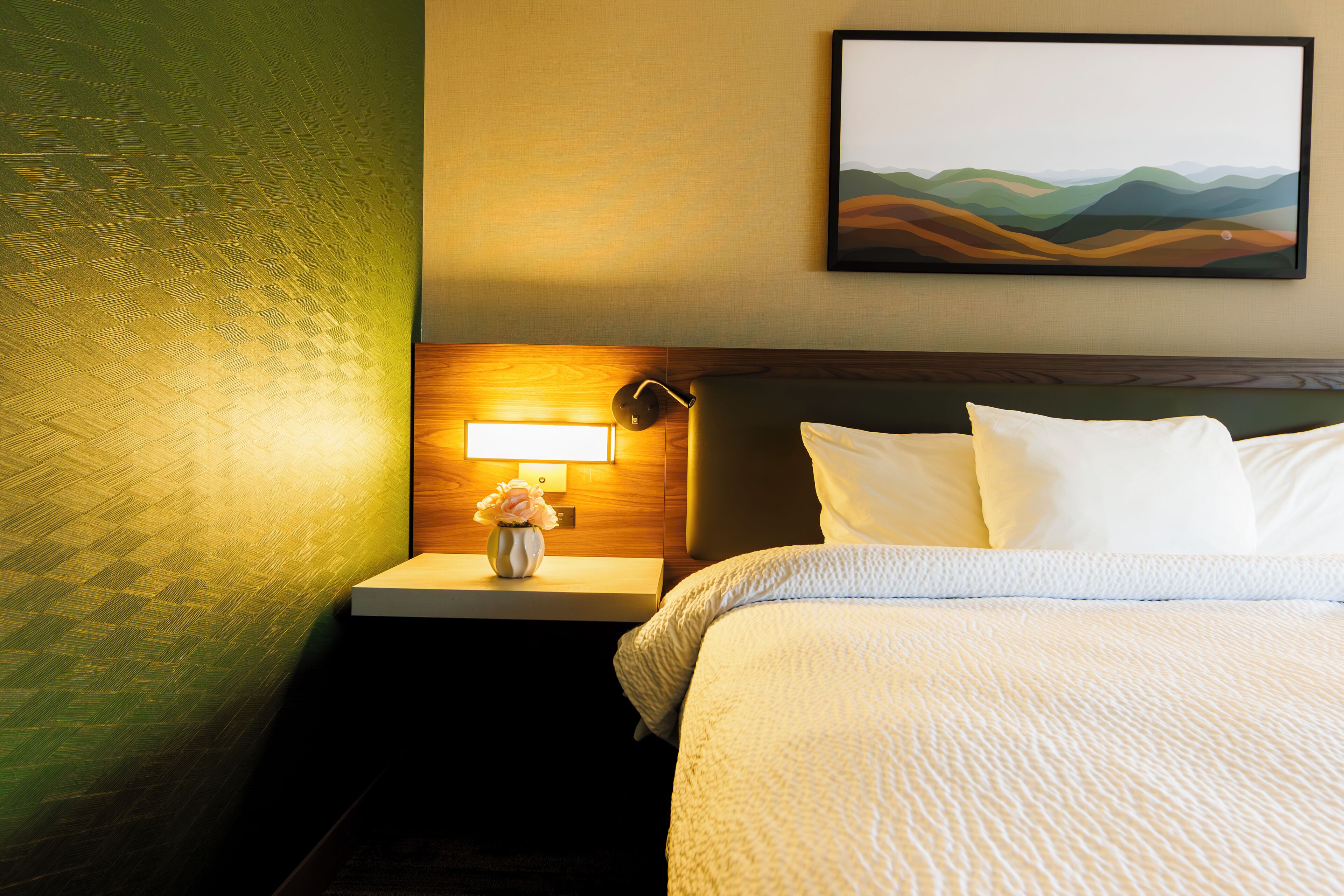 A hotel room with a neatly made bed, a bedside lamp, and a painting on the wall.