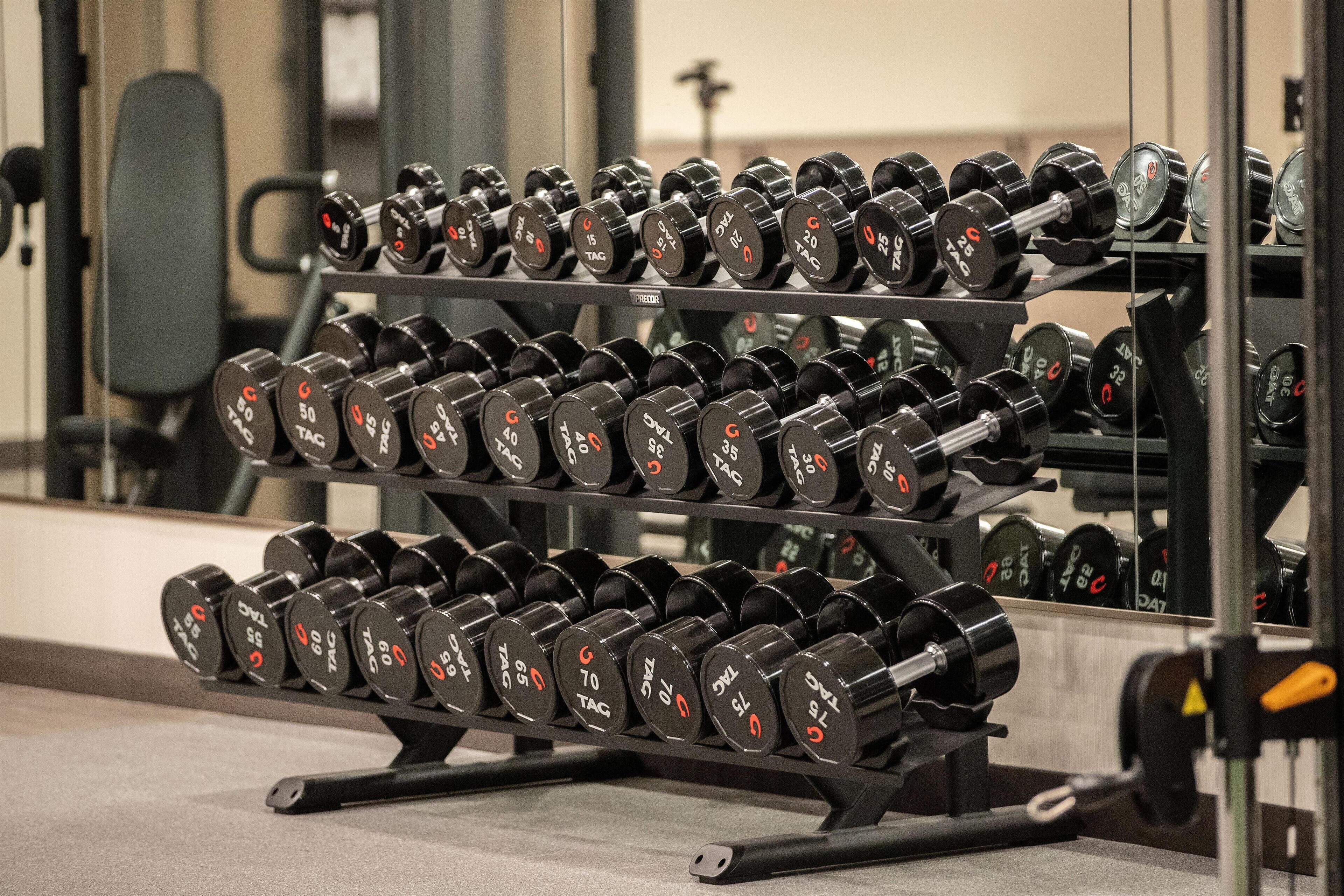 Un estante con mancuernas en un gimnasio, con un espejo que refleja los equipos y una silla al fondo.