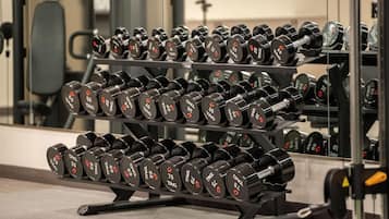 Un estante con mancuernas en un gimnasio, con un espejo que refleja los equipos y una silla al fondo.