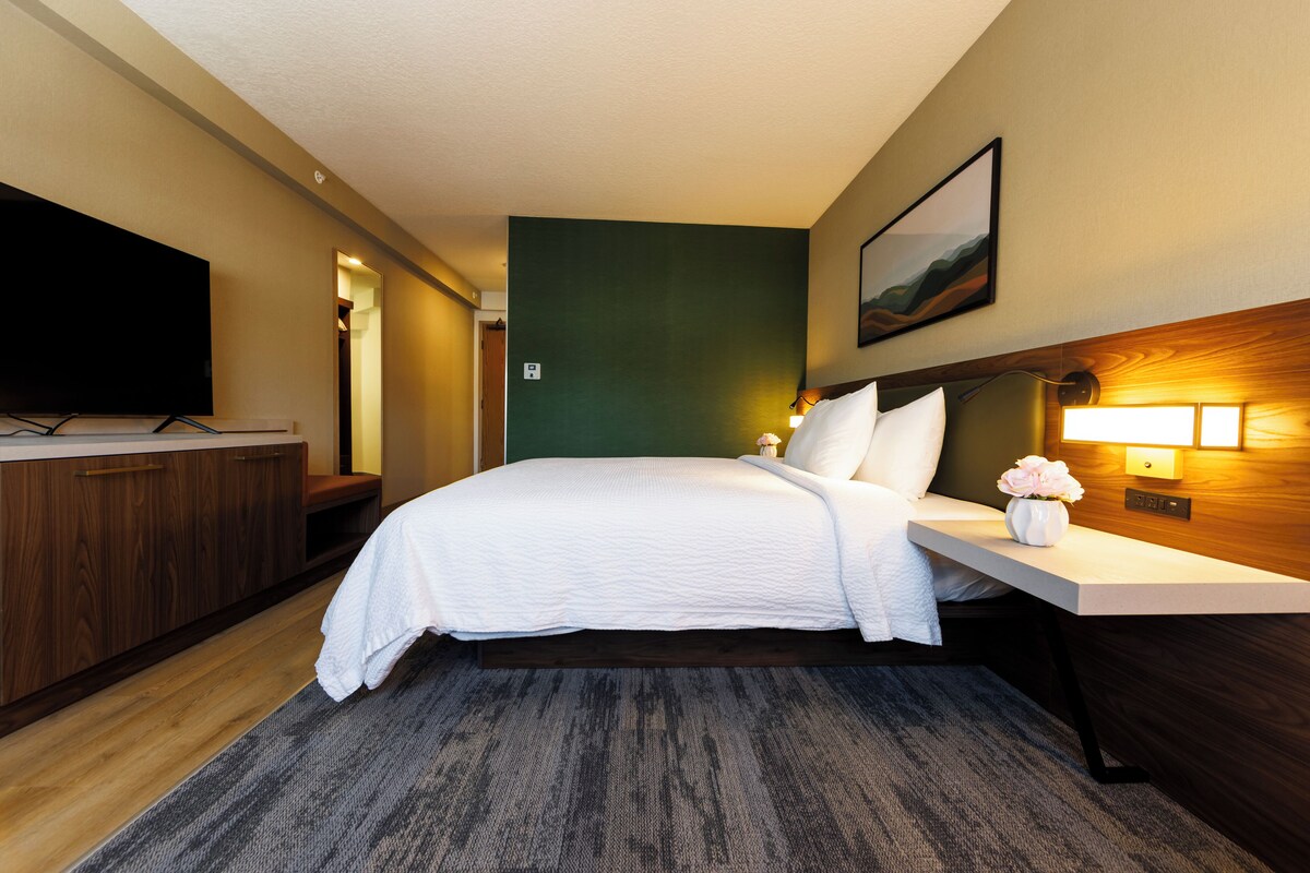A hotel room with a bed, bedside table, lamp, and a flat-screen TV.