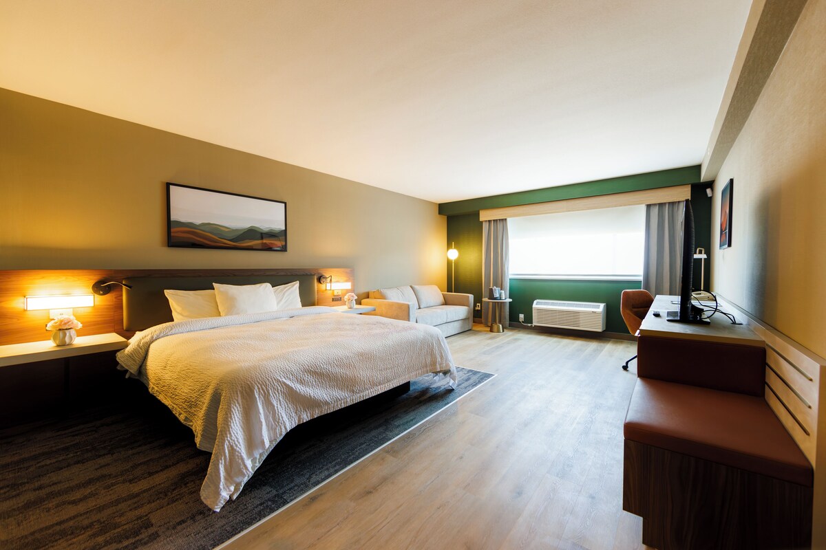 A hotel room with a bed, bedside tables, a desk, a chair, a sofa, and a large window with curtains.
