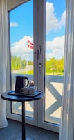 Una habitación con una puerta que da a un balcón, una mesita con una cafetera y tazas, y una bandera en el exterior.