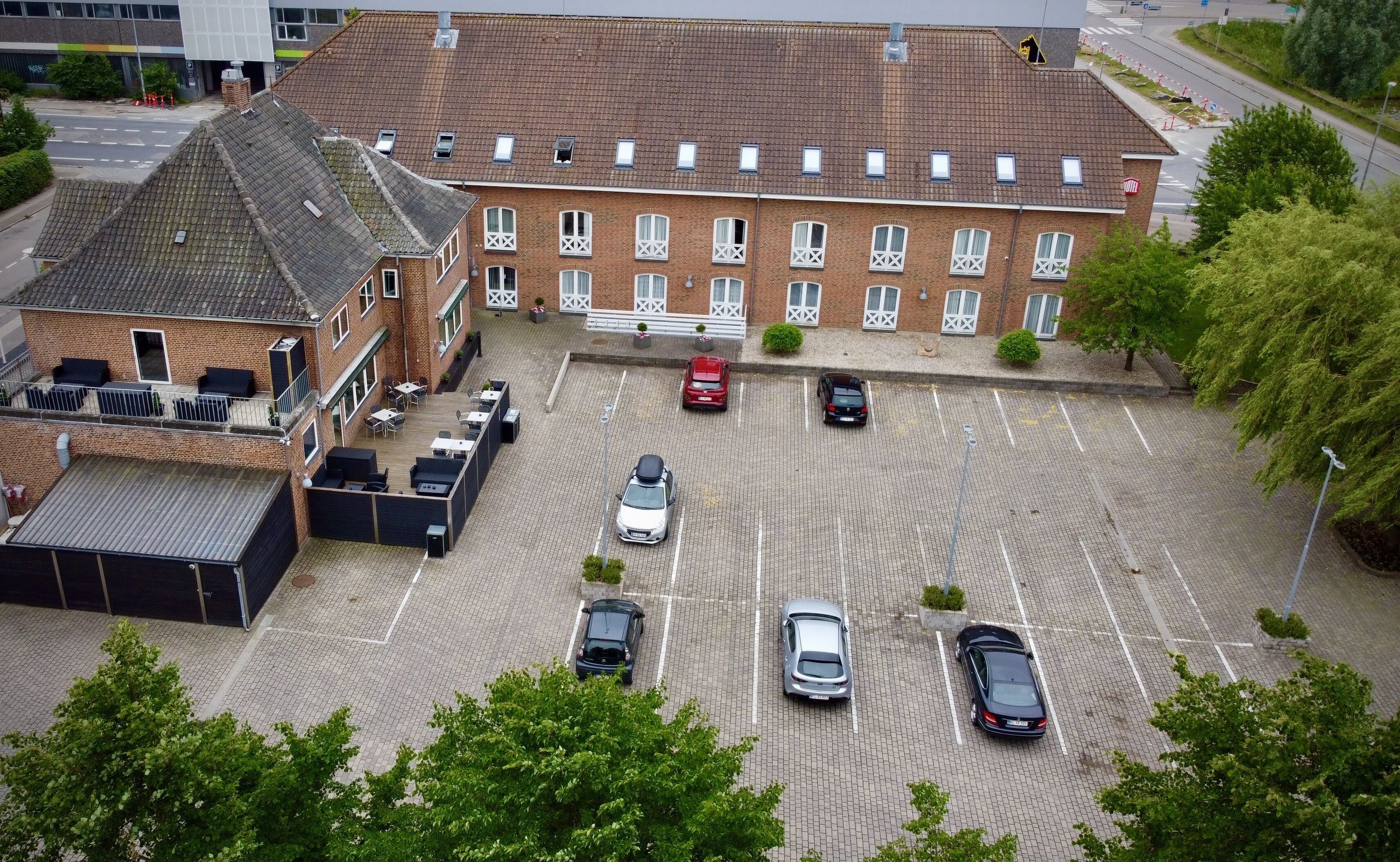 A parking lot with several cars, a building with a brick facade and multiple windows, and a small outdoor seating area with tables and chairs.