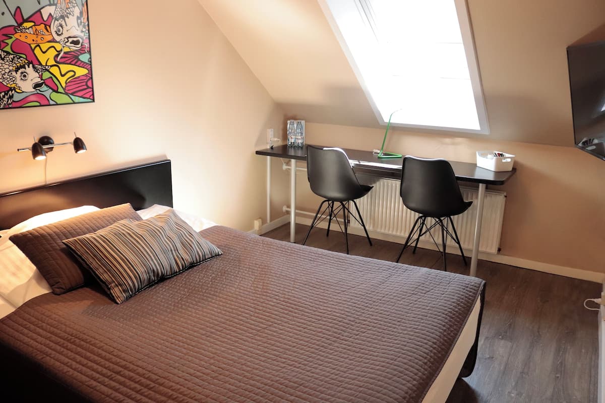 A bedroom with a large bed, a desk with two chairs, a radiator, and a window.