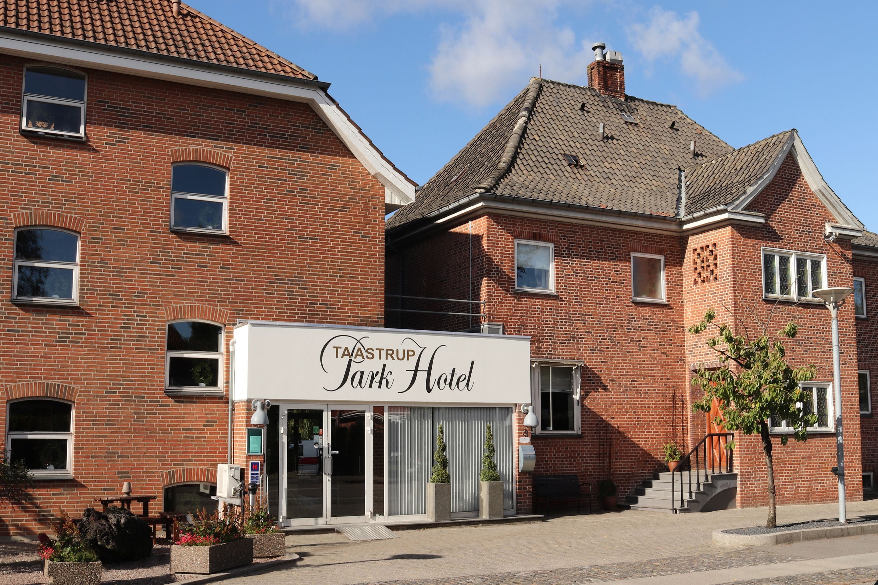 A brick building with a sign for "Taastrup Park Hotel" featuring a modern entrance with glass doors and potted plants.