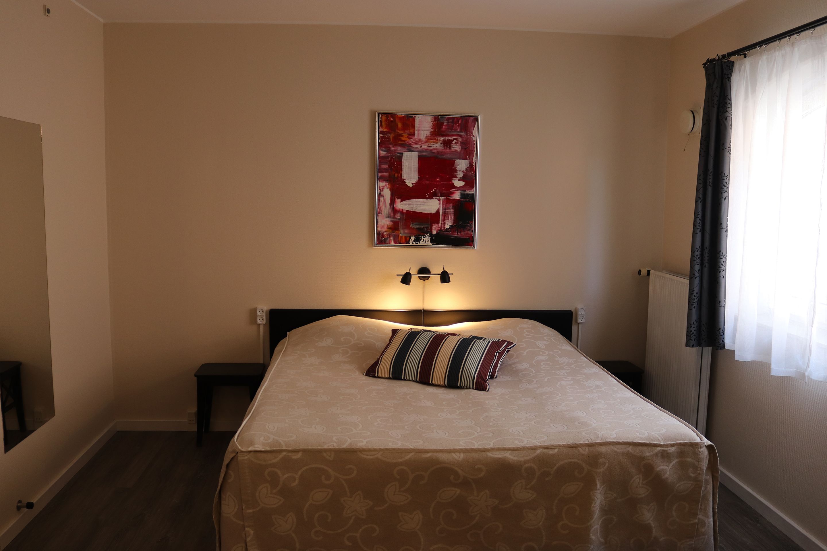 A bedroom with a bed, a nightstand, a lamp, a wall-mounted artwork, and a window with curtains.