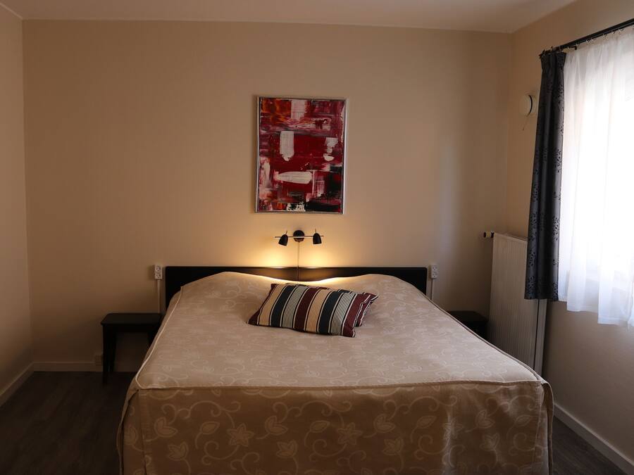 A bedroom with a bed, a nightstand, a lamp, a wall-mounted artwork, and a window with curtains.