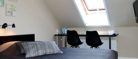 A bedroom with a bed, a desk, two chairs, a skylight, and a wall with travel-themed posters.