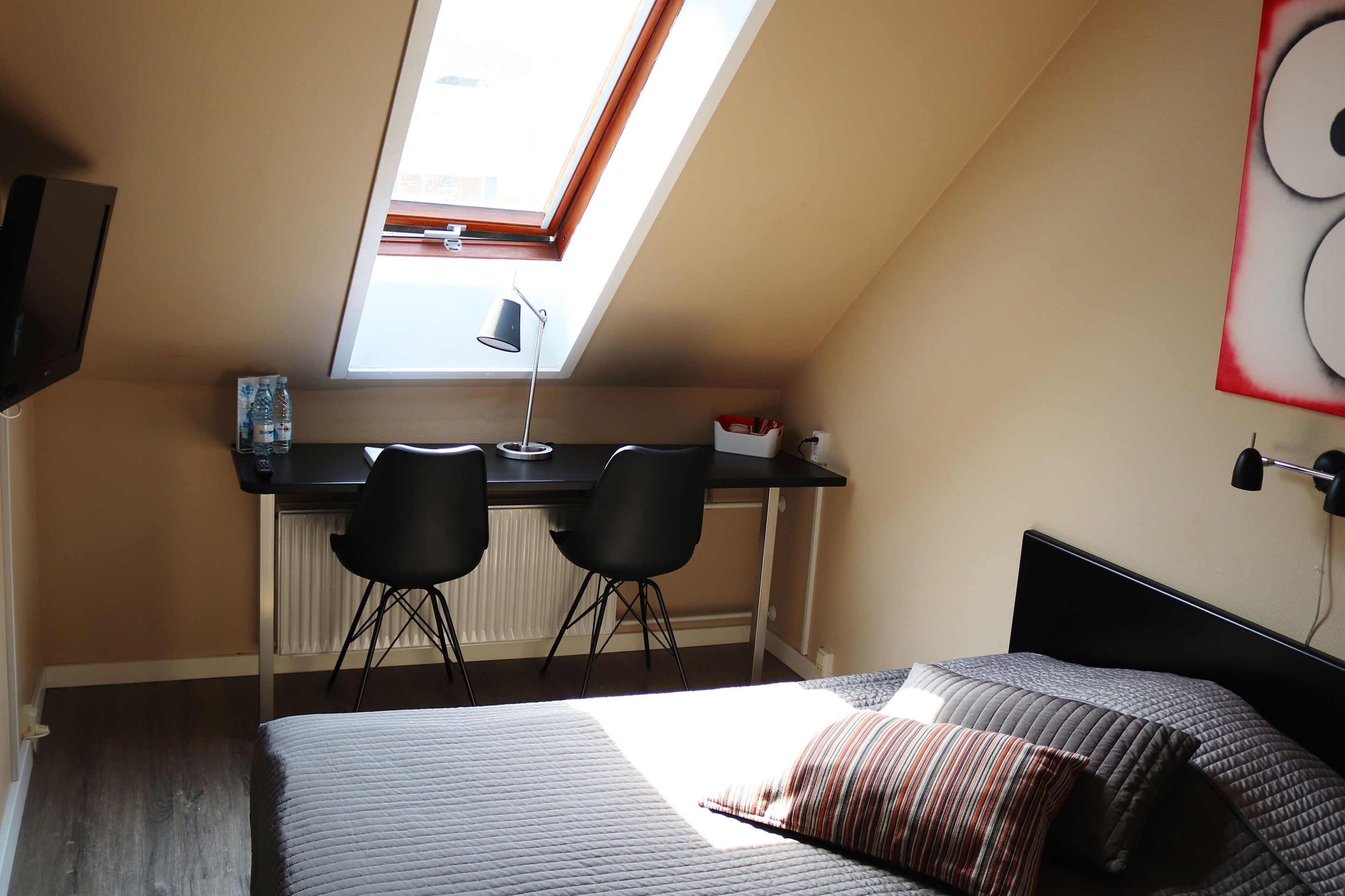 Une chambre à coucher comprenant un lit, un bureau, deux chaises, un puits de lumière et un téléviseur fixé au mur.