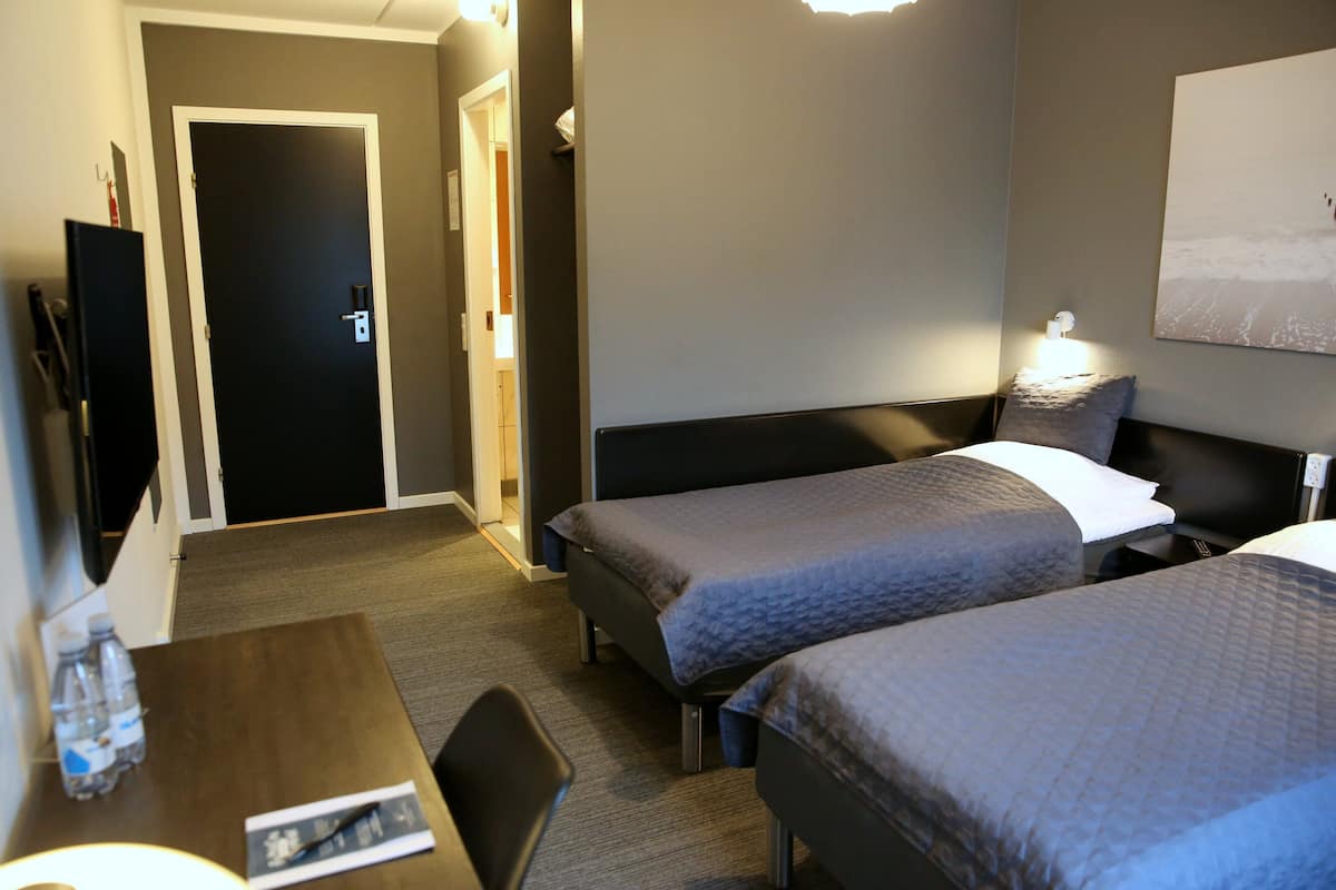 A hotel room with two single beds, a desk with a chair, a TV, and a wall-mounted lamp.
