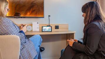 A modern room with a flat-screen TV mounted on the wall, a small table with a laptop and a game controller, and a comfortable seating area with a couch and chairs.