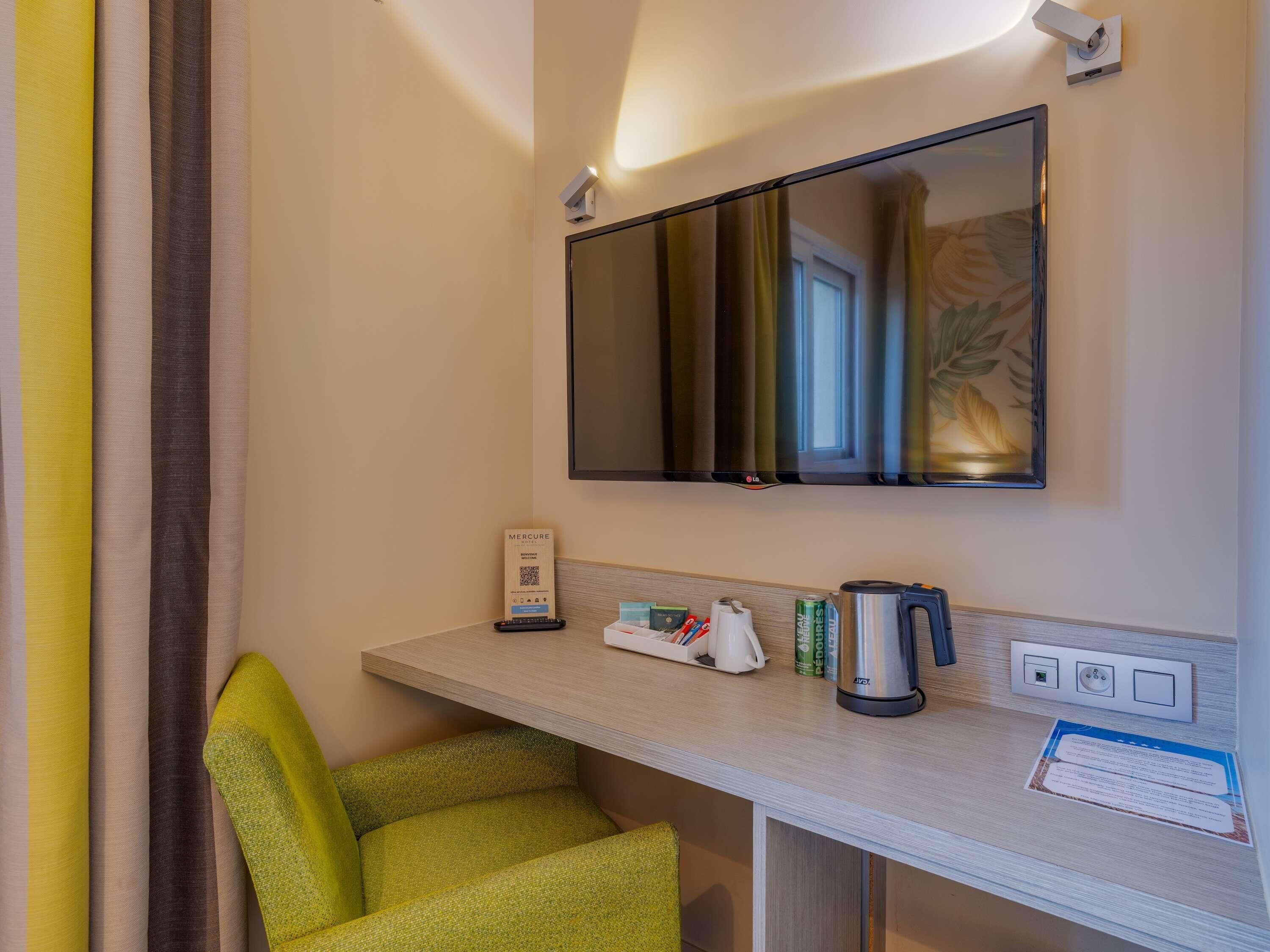 Ein Hotelzimmer mit einem einzelnen grünen Sessel, einem flachbildfernseher an der Wand, einem kleinen Tisch mit Kaffeemaschine und einem wandmontierten Thermostat.