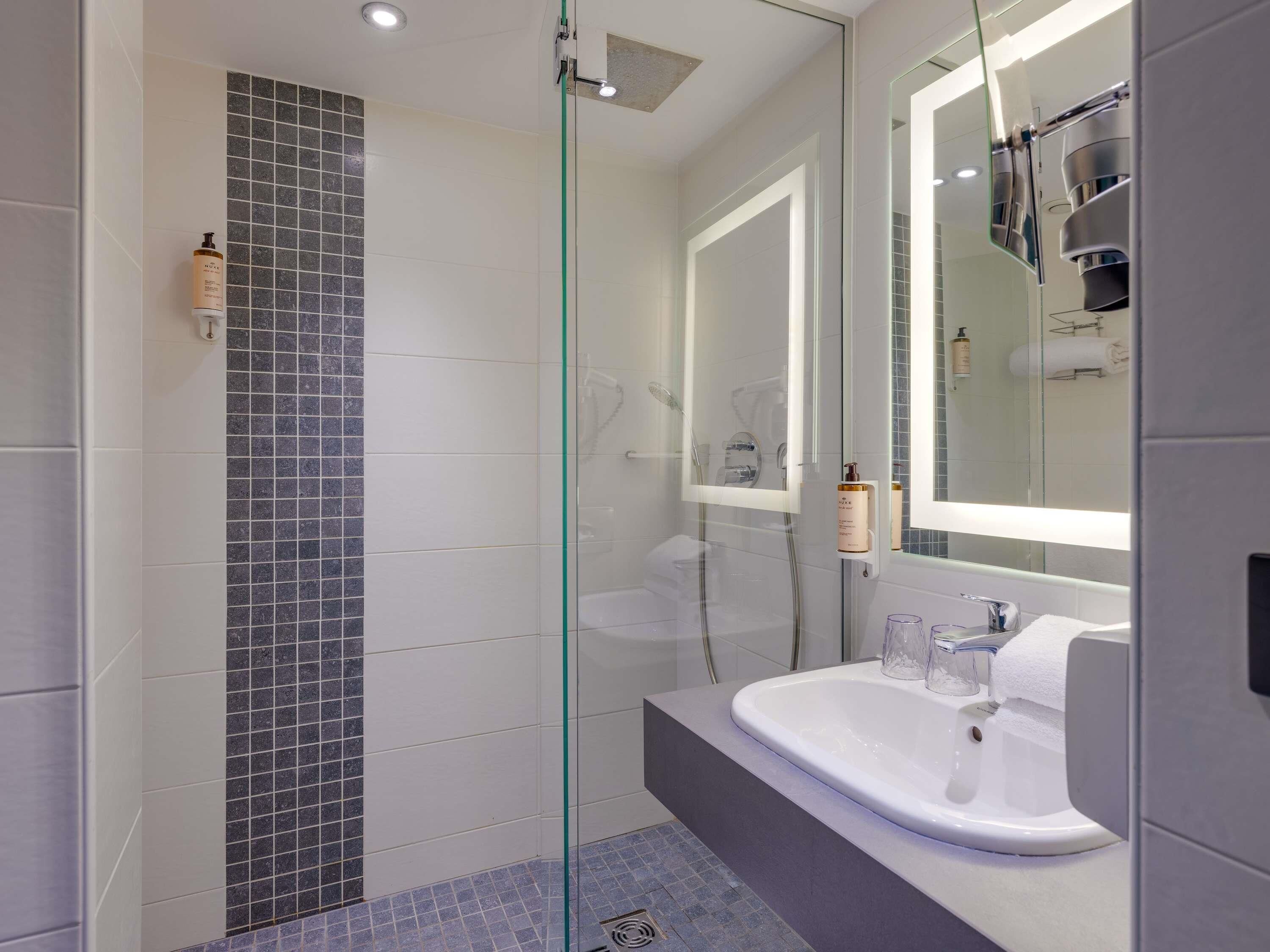 Un baño moderno con ducha de vidrio, lavamanos blanco con espejo y piso de baldosas.