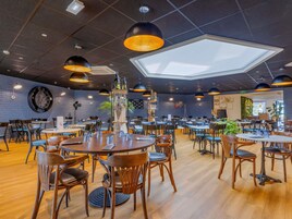 Um restaurante espaçoso com várias mesas e cadeiras, um lustre grande no centro e um relógio na parede.