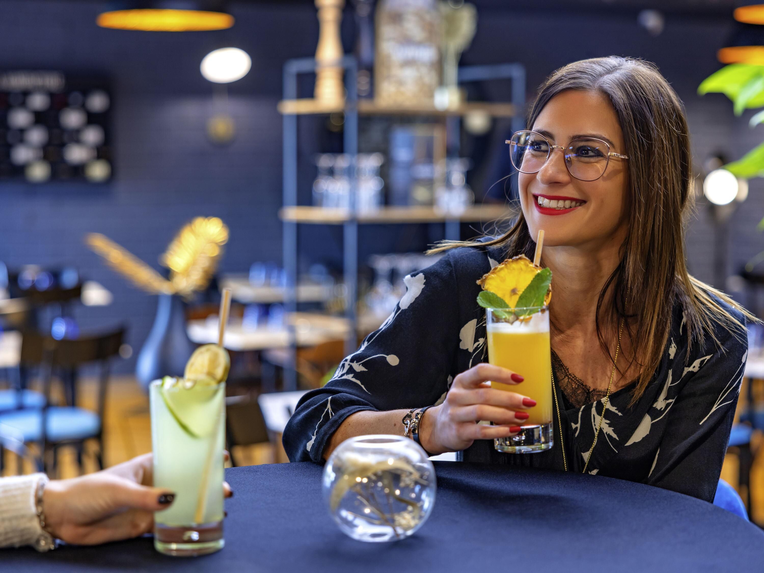 A person holding a drink with a garnish, seated at a table with another drink and a glass.