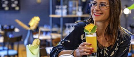 A person holding a drink with a garnish, seated at a table with another drink and a glass.