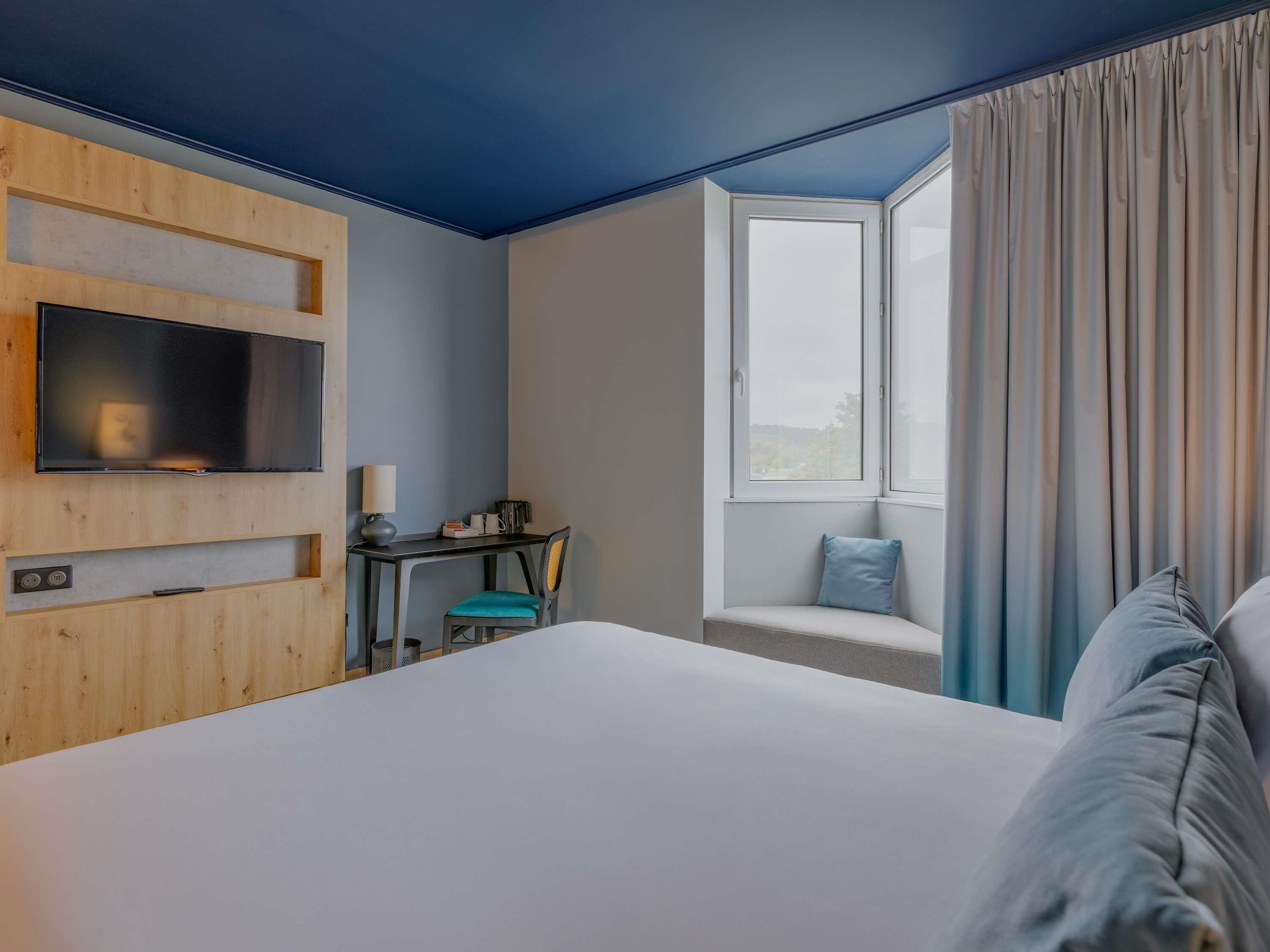 Una habitación de hotel con cama, escritorio, silla, televisor y ventana con cortinas.