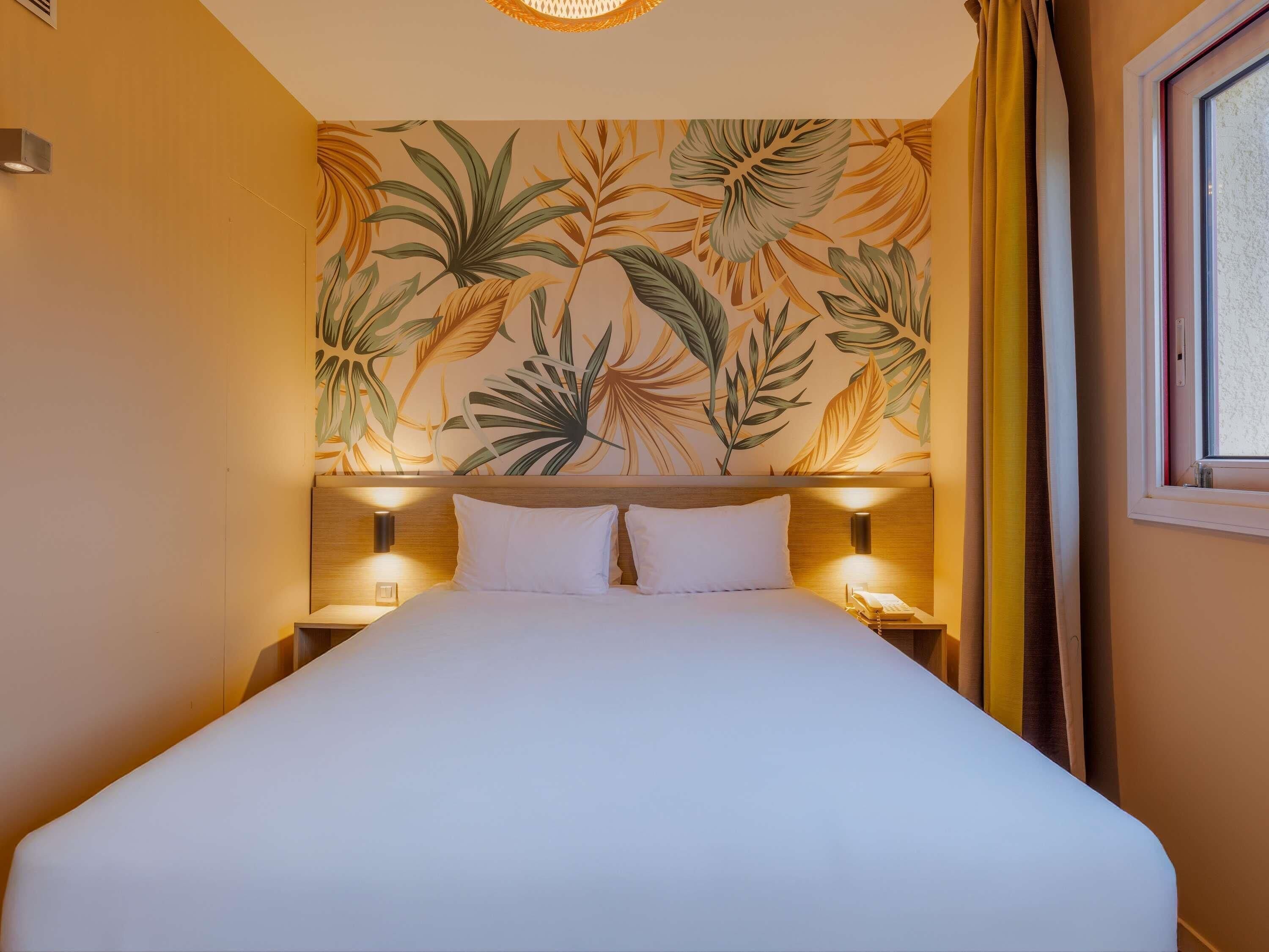 Una camera d'albergo con un letto, due abat-jour, un muro decorato con un motivo a foglia tropicale e una finestra con tende.
