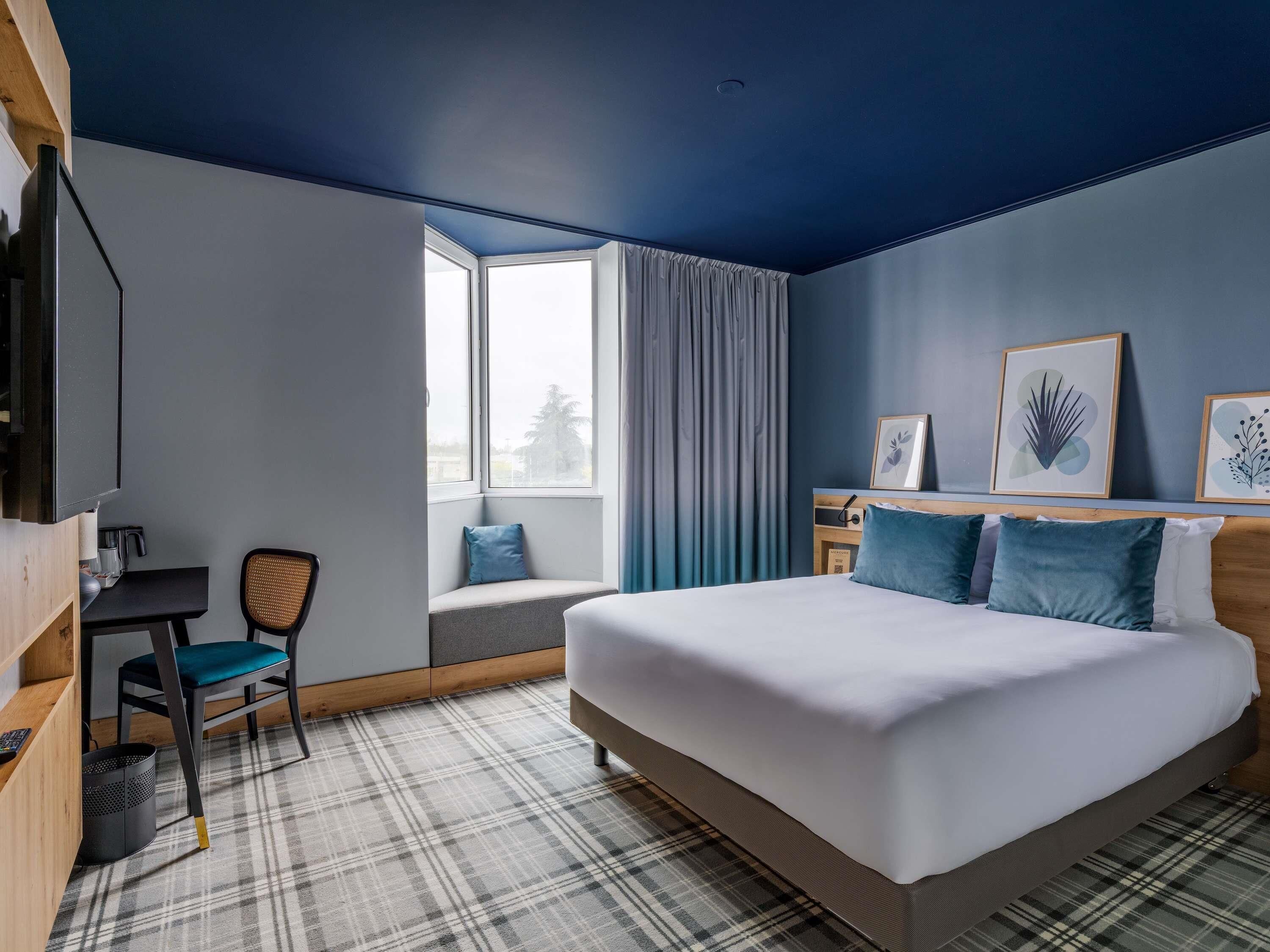 Une chambre d’hôtel avec un lit, un bureau, une chaise et une fenêtre avec des rideaux.