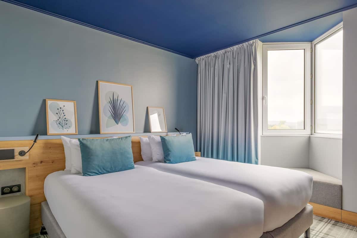 A hotel room with two beds, a window with curtains, and framed artwork on the wall.