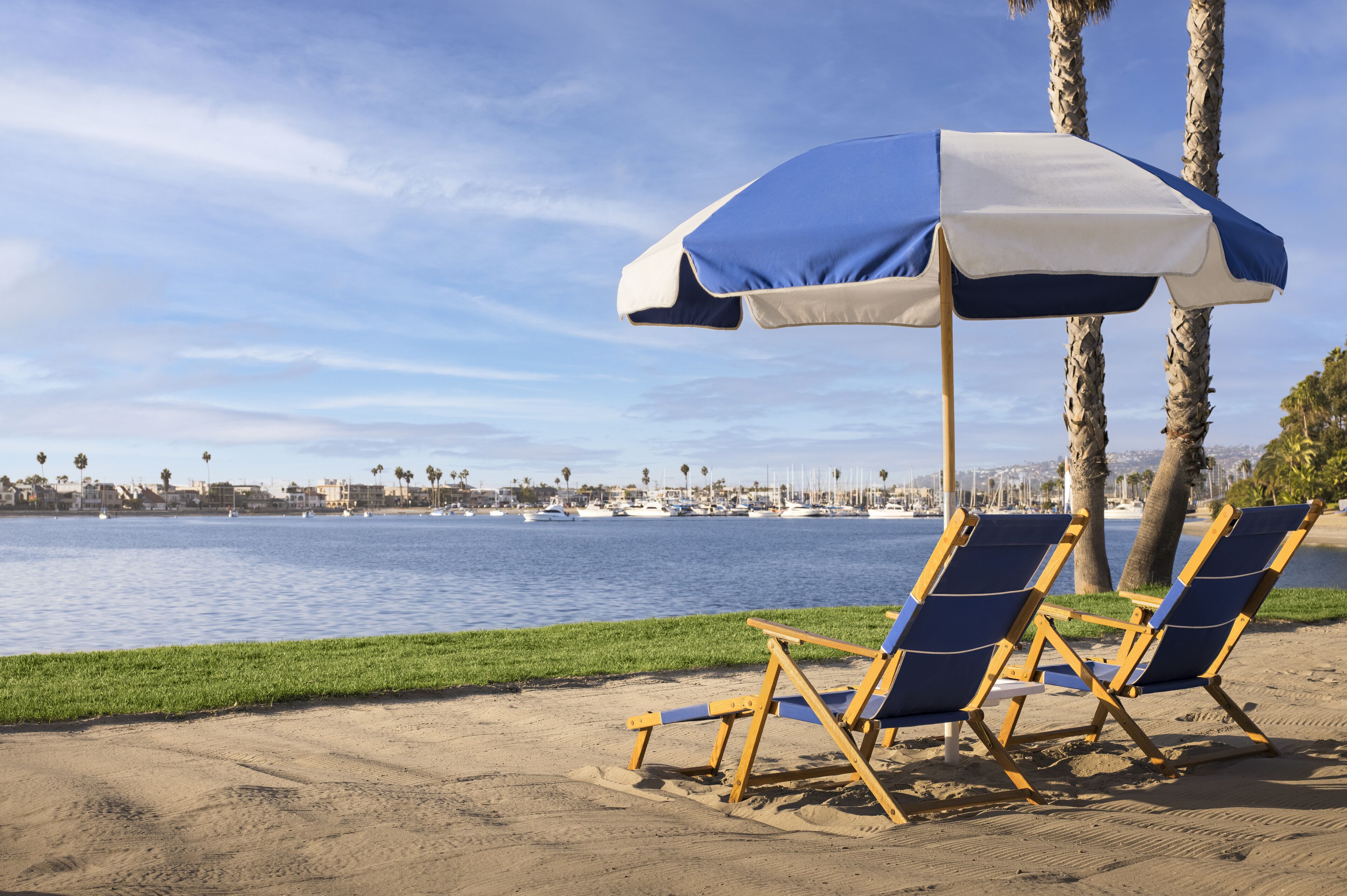 A beach with two blue and white beach chairs under a matching umbrella, a sandy shore, and a marina with boats in the background.