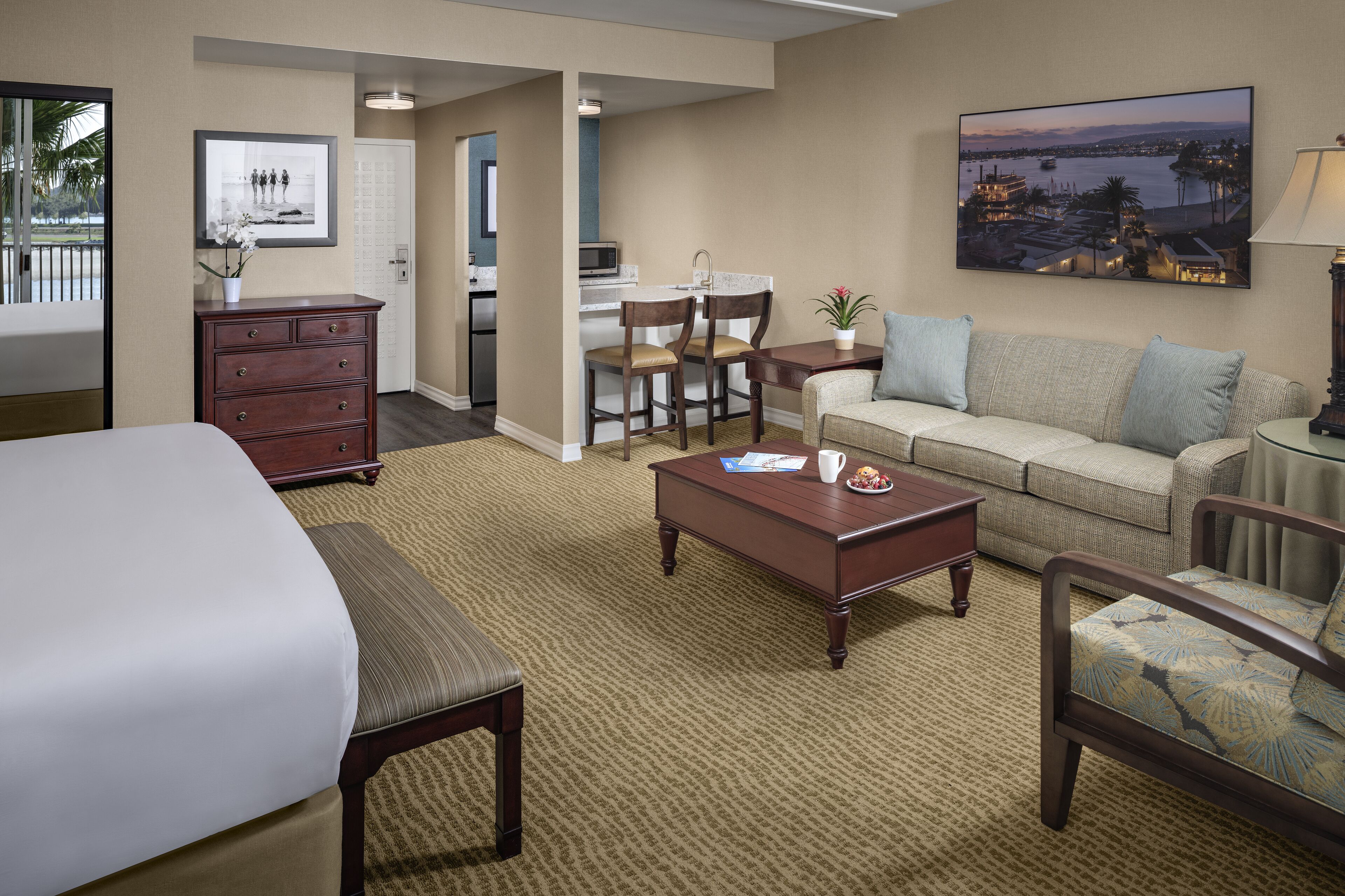 A hotel room with a bed, a sofa, a coffee table, a desk, and a chair. There is a painting on the wall and a plant on the desk.