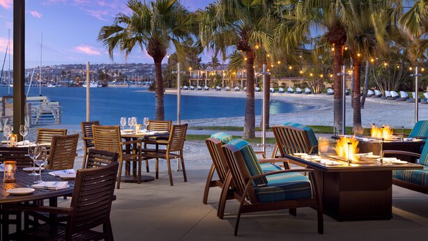 An outdoor dining area with tables and chairs, overlooking a beach and ocean. - Bahia Resort Hotel (San Diego)