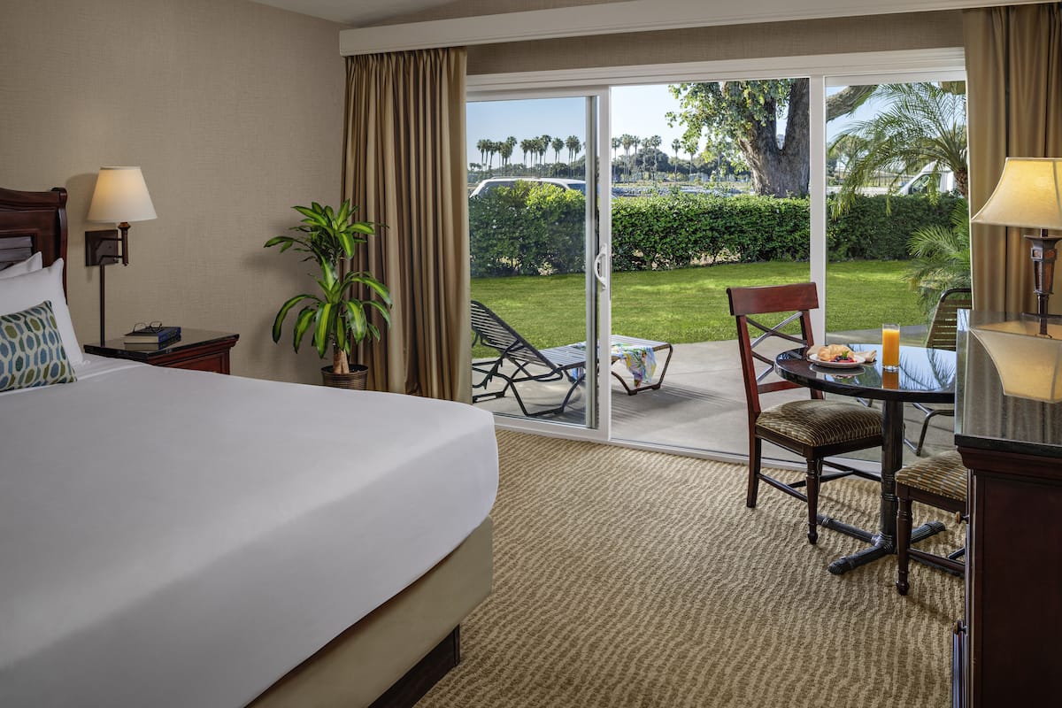 Une chambre d’hôtel avec un grand lit, un bureau avec une chaise, une table avec une chaise, une plante en pot et une vue sur un jardin et des palmiers, visible à travers la porte-fenêtre coulissante.