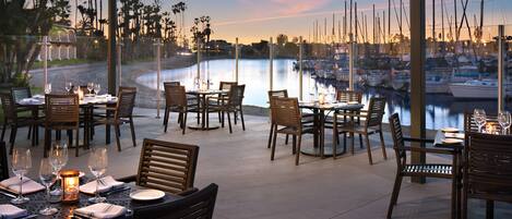 An outdoor dining area with tables and chairs, overlooking a marina with boats and a calm water body.