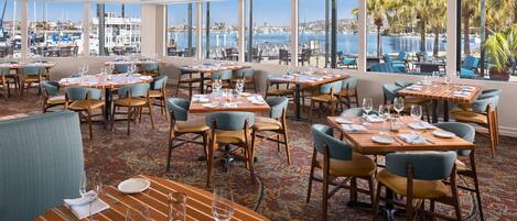 Un restaurante con mesas dispuestas para comer, amplias ventanas que ofrecen vista al puerto deportivo y al agua.
